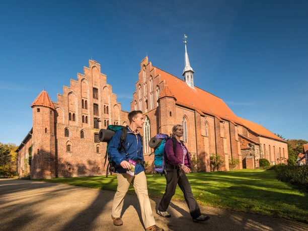 wienhausen_herbst_dk_l-neburger-heide-2015-378-l-neburger-heide-gmbh Zwei Wanderer mit Rucksäcken spazieren entlang einer beeindruckenden Backsteinkirche im Sonnenlicht.