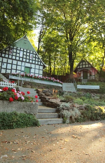 Freilichtbühne Kahle Wart Fachwerkhäuser umgeben von blühenden Blumen und Bäumen auf einer Freilichtbühne bei Sonnenuntergang.