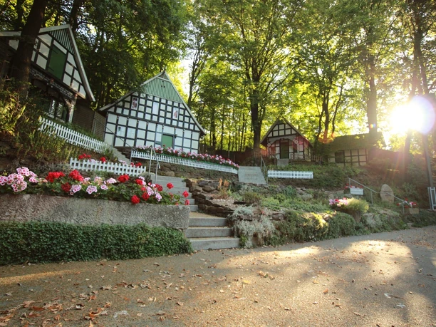 Freilichtbühne Kahle Wart Fachwerkhäuser umgeben von blühenden Blumen und Bäumen auf einer Freilichtbühne bei Sonnenuntergang.