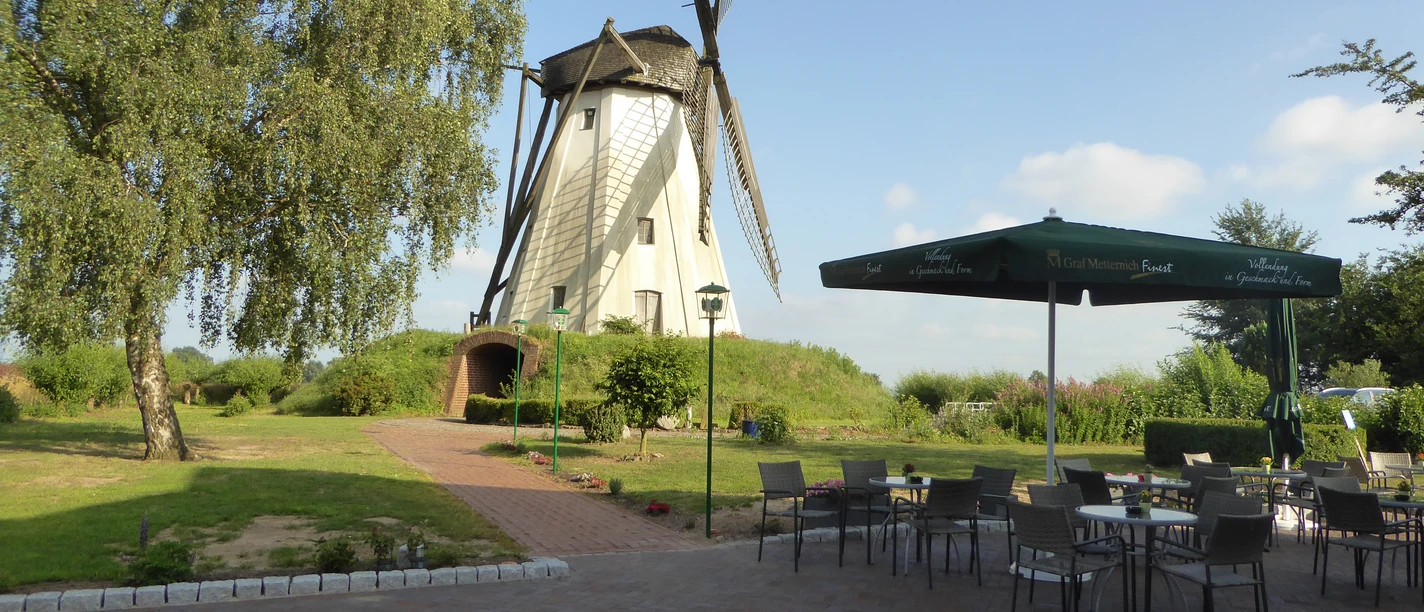 Großenheerser Mühle Mühle mit großen Flügeln, umgeben von grünem Rasen, neben einer Terrasse mit Stühlen und Tischen.