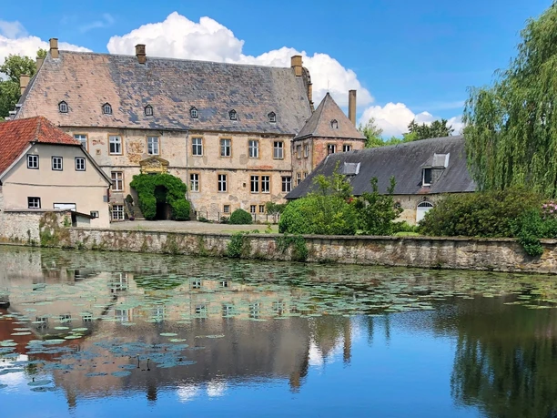 Wasserschloss Tatenhausen Historische Wasserschlossanlage mit malerischem Teich, umgeben von Bäumen und blühenden Seerosen.