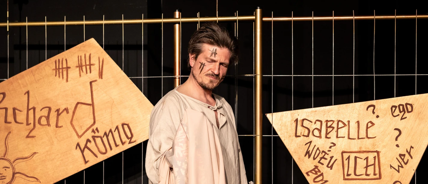 Richard II: A King's Solo An actor playing Richard II in front of a stage curtain and labeled wooden signs