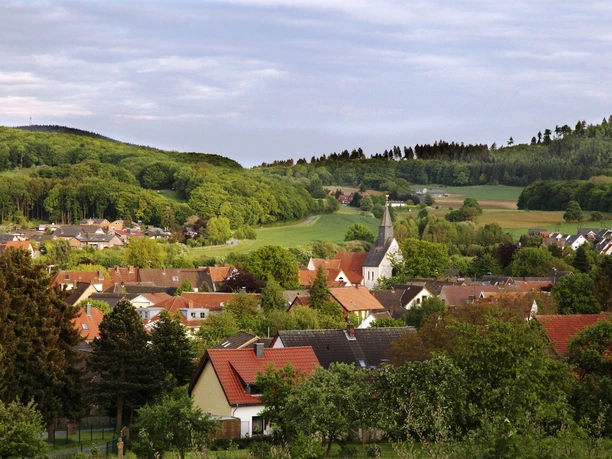 Borgholzhausen Ein idyllisches Dorf mit Kirchturm, umgeben von grünen Feldern und bewaldeten Hügeln im Hintergrund.