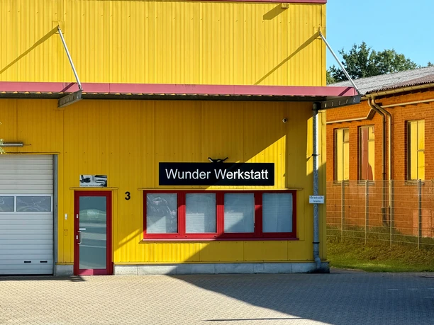 Wunder Werkstatt.jpg Gelbe Werkstattfassade mit Schild „Wunder Werkstatt“, roter Tür und großen Fenstern.