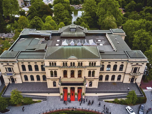 Zoo Gesellschaftshaus Frankfurt_Luftaufnahme des Zoo Gesellschaftshauses Frankfurt Luftaufnahme des Zoo Gesellschaftshauses Frankfurt mit historischem Gebäude, begrüntem Umfeld des Zoos und Veranstaltungseingang mit rotem Teppich. Vor dem Gebäude sind mehrere Personen auf dem Vorplatz zu sehen.
