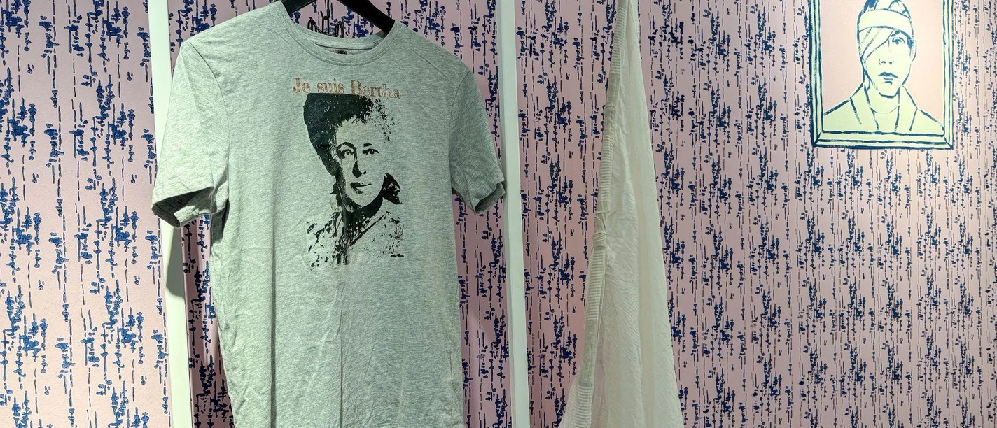 Ein graues T-Shirt mit einem Porträtdruck hängt an einem weißen Kleiderständer. Im Hintergrund ist eine gemusterte Wand und ein weiteres Kunstwerk zu sehen.