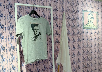Die Waffen nieder Ein graues T-Shirt mit einem Porträtdruck hängt an einem weißen Kleiderständer. Im Hintergrund ist eine gemusterte Wand und ein weiteres Kunstwerk zu sehen.