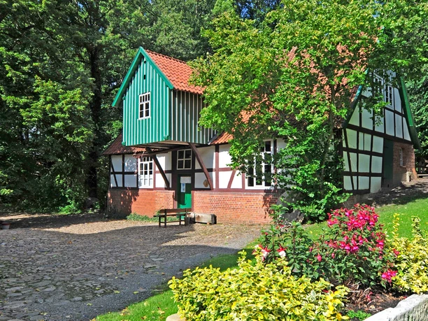 Historische Wassermühle mit roten Ziegelsteinmauern, Fachwerk und grünem Schindeldach im Grünen.