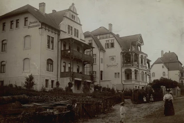 Eine historische Straßenszene mit mehreren Wohnhäusern im klassischen Stil. Menschen stehen im Vordergrund und eine kleine Gruppe ist in der Nähe zu sehen.