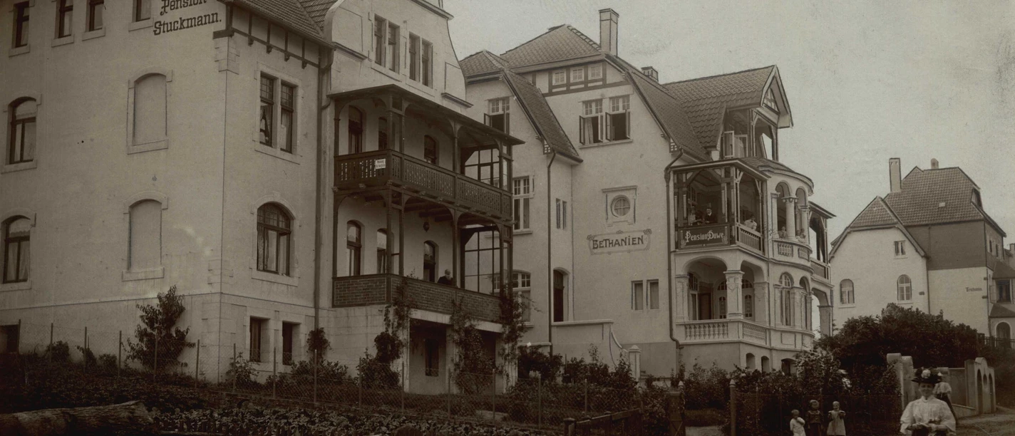 Eine historische Straßenszene mit mehreren Wohnhäusern im klassischen Stil. Menschen stehen im Vordergrund und eine kleine Gruppe ist in der Nähe zu sehen.