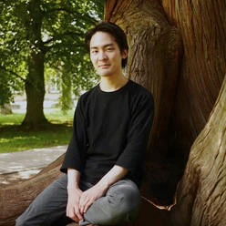 Ein junger Mann sitzt entspannt an einem großen Baum. Im Hintergrund sind grüne Bäume und ein klarer Himmel zu sehen. Es ist der Pianist Teppei Kuroda.