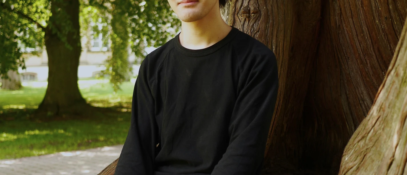 Ein junger Mann sitzt entspannt an einem großen Baum. Im Hintergrund sind grüne Bäume und ein klarer Himmel zu sehen. Es ist der Pianist Teppei Kuroda.