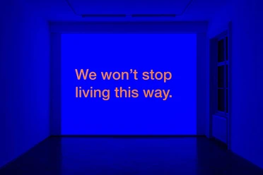 We Won't Stop Living This Way - Felix Gaudlitz An exhibition space illuminated in blue with orange lettering projected onto the wall