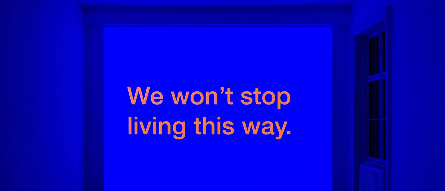 We Won't Stop Living This Way - Felix Gaudlitz An exhibition space illuminated in blue with orange lettering projected onto the wall