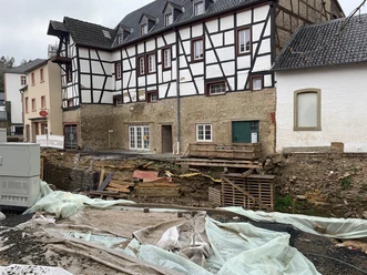 20.10._Hochwasser2 Ein historisches Gebäude mit weißem Fachwerk und grauem Dach steht neben einer Baustelle. Der Boden ist aufgerissen, und Baustellenmaterialien sind überall verteilt.