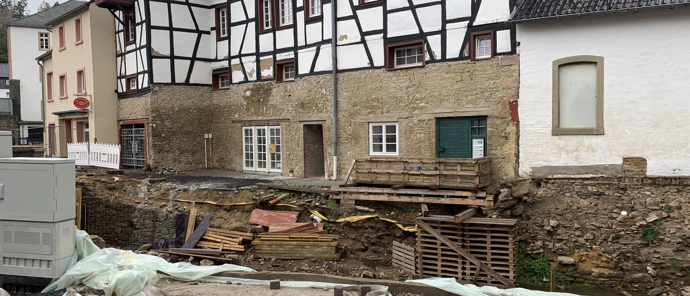 20.10._Hochwasser2 Ein historisches Gebäude mit weißem Fachwerk und grauem Dach steht neben einer Baustelle. Der Boden ist aufgerissen, und Baustellenmaterialien sind überall verteilt.