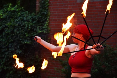 Feuershow 1.jpg Feuerkünstlerin in Rot mit brennenden Fackeln vor einer Backsteinmauer.