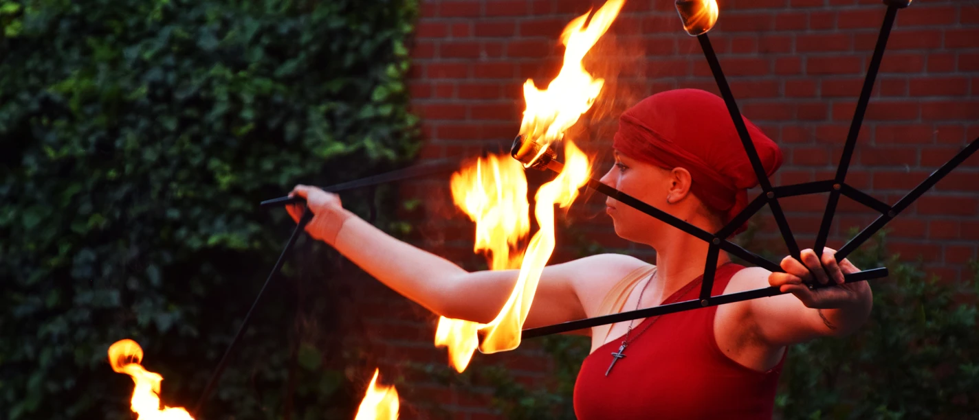 Feuershow 1.jpg Feuerkünstlerin in Rot mit brennenden Fackeln vor einer Backsteinmauer.