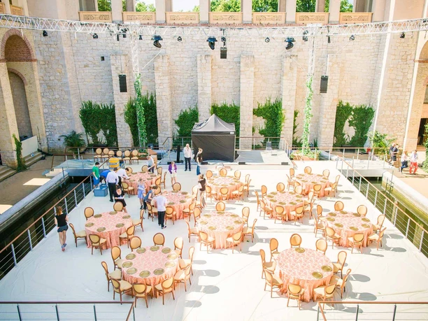 BALZER & Co. GmbH_Event Catering_Außenbereich einer Eventlocation Außenbereich einer Eventlocation mit gedeckten Rundtischen, Stühlen, Bühne und Veranstaltungstechnik. Mehrere Personen bereiten die Fläche für ein Catering oder eine Veranstaltung vor.