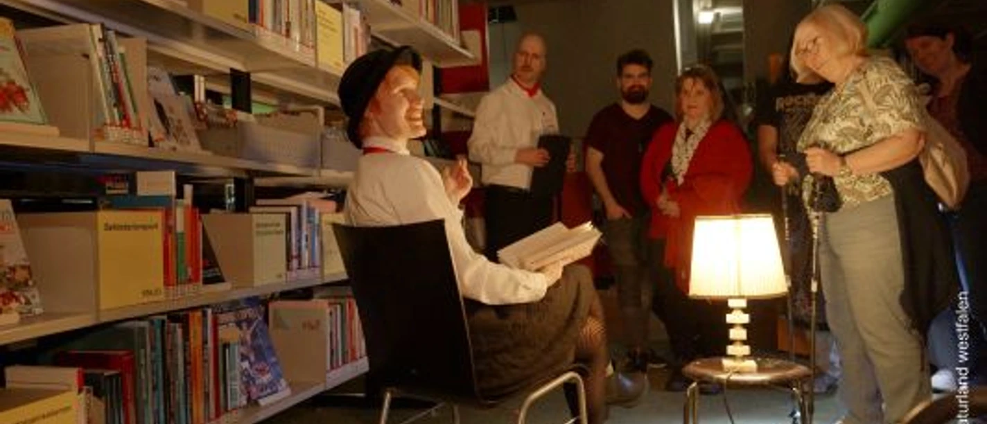 Szenenfoto: ein Mann sitzt vor einem Bücherregal, Publikum interagiert