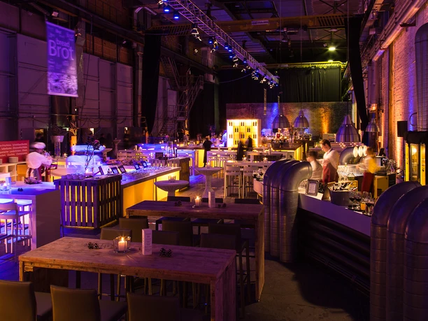 BALZER & Co. GmbH_MICE Agentur_Dunkler Veranstaltungssaal Dunkler Veranstaltungssaal mit Bühne, warmem Bühnenlicht und einem langen gedeckten Tisch für ein Gala-Dinner. Im Vordergrund sind weitere gedeckte Rundtische mit Kerzen und Dekoration zu sehen.