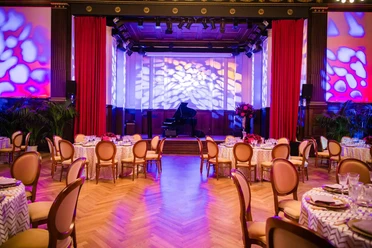 BALZER & Co. GmbH_MICE Agency_Festively Decorated Event Hall Festlich eingerichteter Veranstaltungssaal mit gedeckten Rundtischen, Bühne, Flügel, roten Vorhängen und farbiger Lichtinszenierung. Der Raum ist für ein Abendessen oder eine MICE-Veranstaltung vorbereitet.