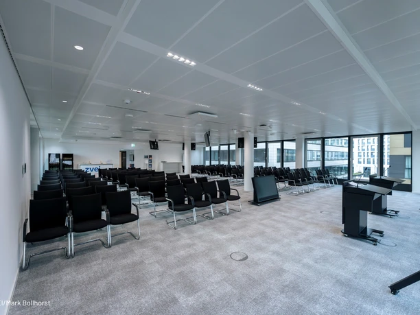 ZVEI-Services GmbH_Siemens Heller Tagungsraum „Siemens“ der ZVEI-Services GmbH mit Reihenbestuhlung, Rednerpult, Monitoren und Präsentationstechnik. Große Fenster geben den Blick auf umliegende Gebäude frei.