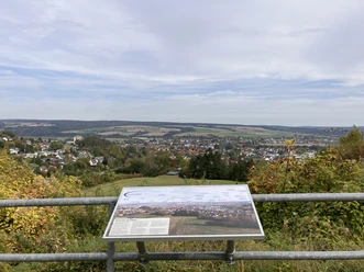 Eine Informationstafel hängt an einem Geländer und zeigt die Panorama-Aussicht des Standortes.