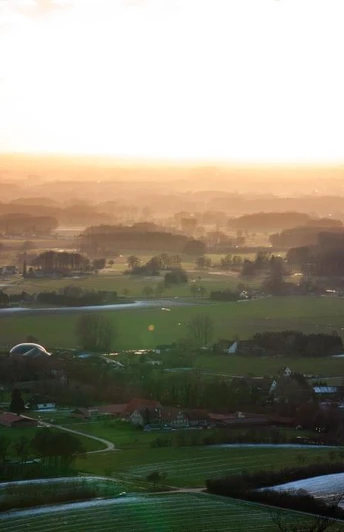 Ländliche Landschaft im Abendlicht, mit Feldern, Wäldern und Häusern unter einem goldenen Himmel.