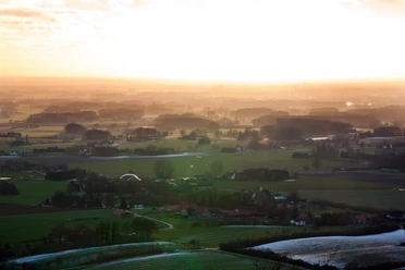 Panoramablick Ländliche Landschaft im Abendlicht, mit Feldern, Wäldern und Häusern unter einem goldenen Himmel.