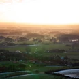 Panoramablick Ländliche Landschaft im Abendlicht, mit Feldern, Wäldern und Häusern unter einem goldenen Himmel.