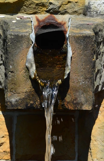 Alte steinerne Wasserquelle mit rostigem Auslaufrohr an einer weißen und gelben Steinwand.