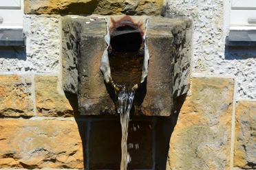 Quellwasser Alte steinerne Wasserquelle mit rostigem Auslaufrohr an einer weißen und gelben Steinwand.