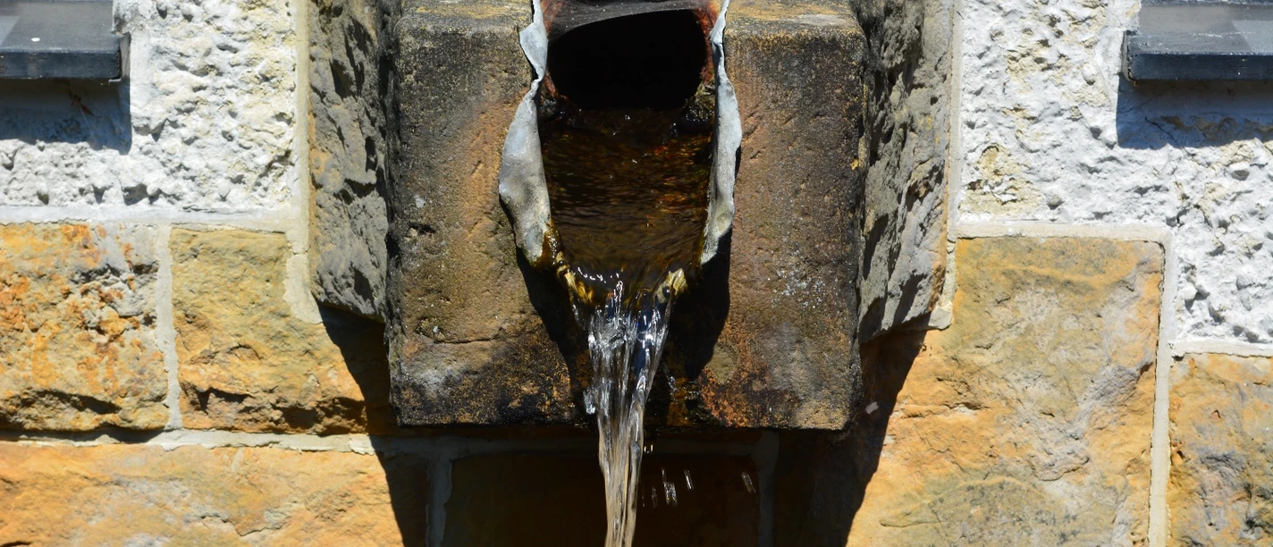 Quellwasser Alte steinerne Wasserquelle mit rostigem Auslaufrohr an einer weißen und gelben Steinwand.