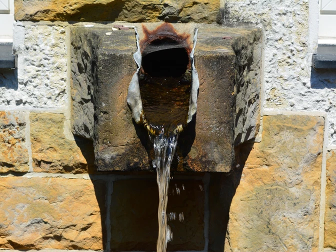 Quellwasser Alte steinerne Wasserquelle mit rostigem Auslaufrohr an einer weißen und gelben Steinwand.
