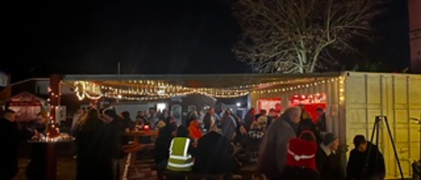 Abendstimmung auf dem Weihnachtsmarkt.jpeg Abendlicher Weihnachtsmarkt in Apen mit Lichterketten, Buden und Besuchergruppen.