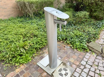 Petershagen Drinking Water Fountain (2).jpeg A metal drinking fountain on a paved path in front of the greenery at Petershagen City Hall.