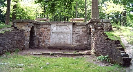 Luccaburg Steinerner Pavillon im Wald, von Bäumen umgeben, mit seitlichen Stufen und zentralem Relief.