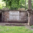 Luccaburg Steinerner Pavillon im Wald, von Bäumen umgeben, mit seitlichen Stufen und zentralem Relief.