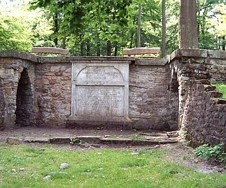 Luccaburg Steinerner Pavillon im Wald, von Bäumen umgeben, mit seitlichen Stufen und zentralem Relief.