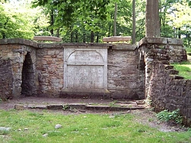 Luccaburg Steinerner Pavillon im Wald, von Bäumen umgeben, mit seitlichen Stufen und zentralem Relief.
