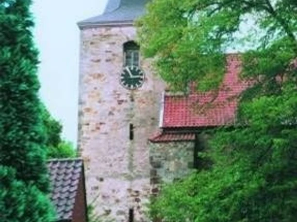 Nendorf Historische Steinkirche mit spitzem Turmdach, grüne Bäume und rote Ziegelbauten im Hintergrund.
