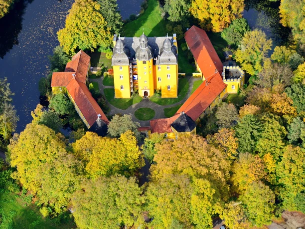 Holter Jagdschloss Luftbild Luftaufnahme des gelben Holter Jagdschlosses mit drei Gebäudeflügeln, umgeben von Herbstbäumen.