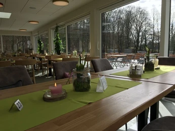 Ein lichtdurchfluteter Wintergarten mit langen Holztischen, Platznummerierung und Blick ins Grüne.