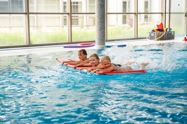 Kinder schwimmen mit Wassermatte Drei Kinder schwimmen fröhlich auf Schwimmbrettern in einem Innenpool. Die ruhige Wasseroberfläche spiegelt das Licht und schafft eine einladende Atmosphäre.