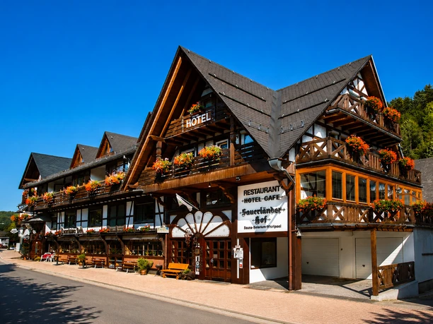 Hausansicht Hotel Sauerländer Hof Willingen Hotel Sauerländer Hof in Fachwerkoptik mit Balkonen, Blumenschmuck, Restaurant-Schild und blauem Himmel in Willingen
