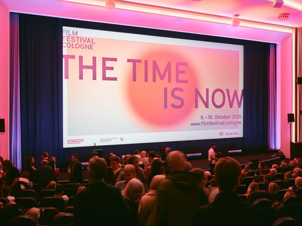 FILM FESTIVAL COLOGNE A movie theater at the Film Festival Cologne, with an audience and a large screen bathed in pink light