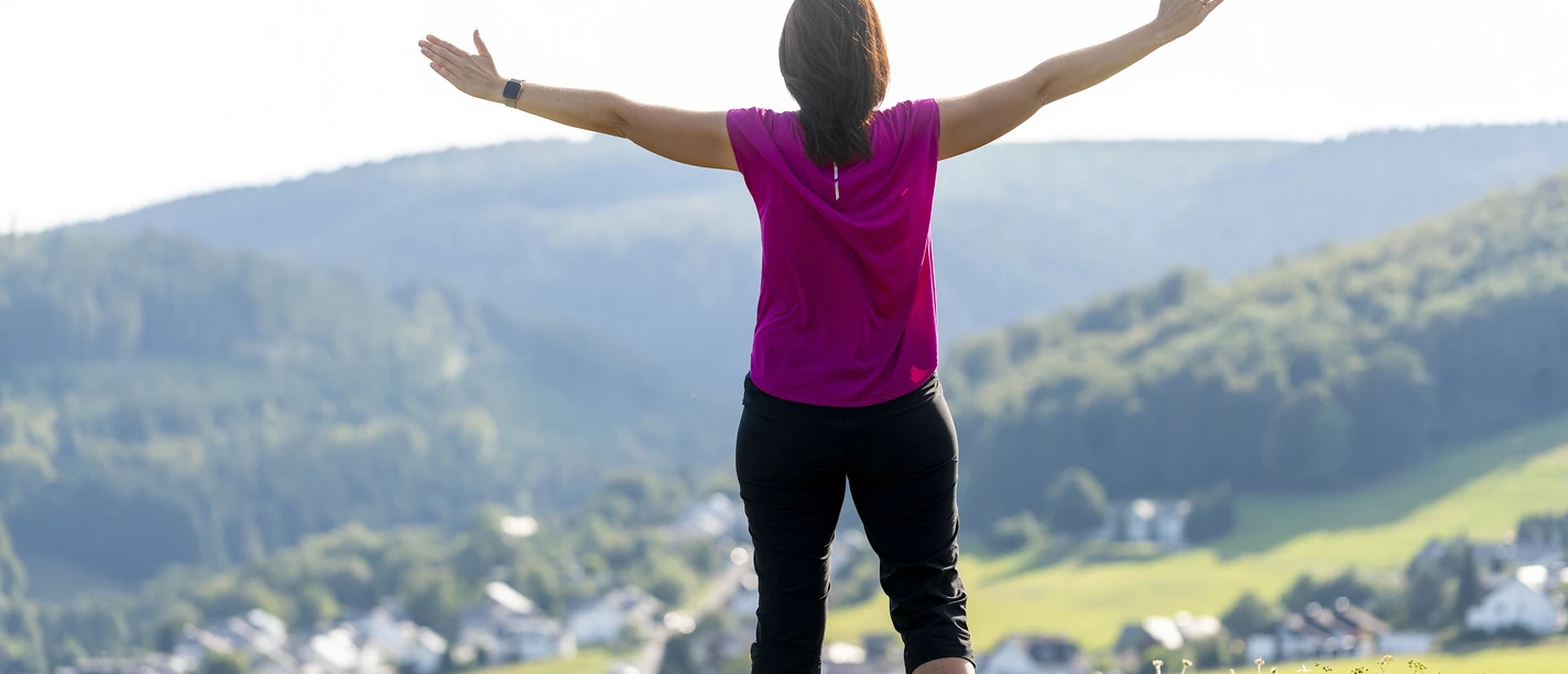 Frau mit ausgebreiteten Armen auf einer Wiese in Willingen Frau mit ausgebreiteten Armen blickt über grüne Hügel und den Ort Willingen im sonnigen Upland.