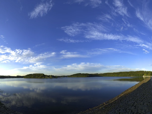 Bevertalsperre Panoramaaufnahme eines großen Sees mit ruhiger Wasseroberfläche und bewaldeten Ufern unter blauem Himmel.