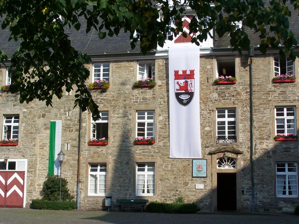 Schloss Hückeswagen Ein historisches Steingebäude mit Blumenkästen, Fahnen und einem gepflasterten Hof im Vordergrund.